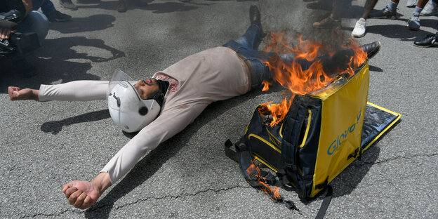 Ein Mitarbeiter des Essenslieferservice Glovo liegt auf der Straße neben einem brennenden Glovo-Rucksack