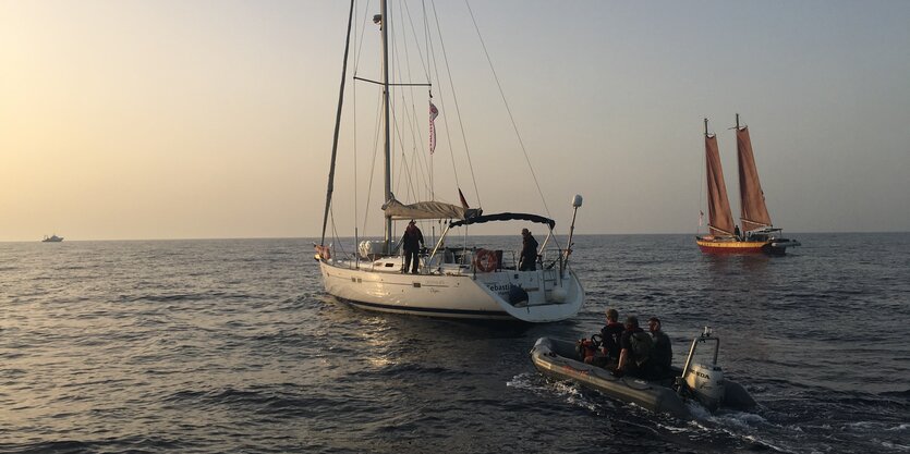 Ein Segelboot segelt in den Sonnenuntergang, dicht gefolgt von einem motorisierten Schlauchboot. Rechts im Bild sieht man ein weiteres Segelboot in der Ferne