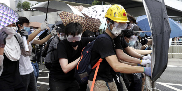 Menschen mit Schirmen, Helmen und Mundschutz stehen hinter einer Absperrung