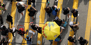 Menschen gehen über eine mit gelben Streifen markierte Straße, einer trägt einen gelben Schirm