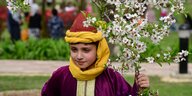 EIn Junge aus der Region Kaschmir in Indien mit traditioneller Kopfbedeckung aus gelber und roter Seide hält mit seiner linken Hand den Ast eines blühenden Mandelbaums fest.