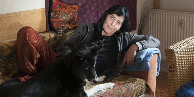 Eine schwarzhaarige Frau sitzt mit einem schwarzen Hund auf einem Sofa und blickt freundlch in die Kamera