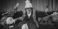 In einem schwarz-weißen Filmstill ist ein grauhaariger Mann mit Mütze zu sehen, der mit mehreren Gegenständen hantiert und außer sich wirkt