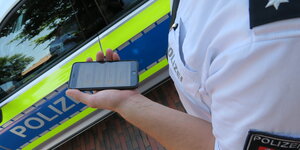 Ein Polizist neben einem Polizeiauto hält ein Smartphone in der Hand