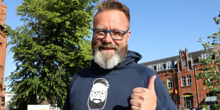 Mitfünziger mit graumelliertem Hipsterbart reckt den Daumen. Auf seinem Sweatshirt: Ein Bild von sich selbst