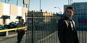 Ein Mann in Anzug steht vor einem Zaun, im Hintergrund ein Polizist