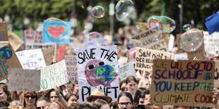 Eine Straße voller Menchen, die Plakate hochhalten, auf denen Klimaparolen stehen. Über den Köpfen der Menge fliegen Seifenblasen.