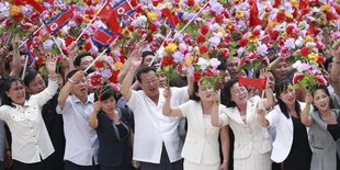 Viele Menschen mit Blumen und roten Fähnchen in der Hand säumen eine Straße.