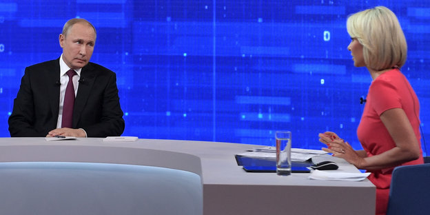Wladimir Putin sitzt in einem blauen Fernsehstudio einer Moderatorin gegenüber