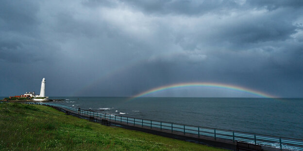 Ein Regenbogen leuchtet zwischen dunklen Wolken hervor, links im Bild ist ein Leuchtturm zu sehen.