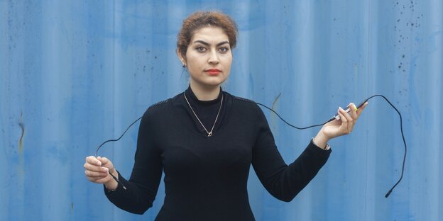 Eine Junge Frau steht vor einer blauen Wand und hält ein Kabel in der Hand