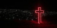Dunkle Nacht, unten leuchtet die Stadt, oben steht ein Kreuz aus rot leuchtenden Konturen