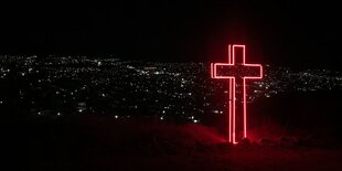 Dunkle Nacht, unten leuchtet die Stadt, oben steht ein Kreuz aus rot leuchtenden Konturen