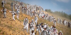 Eine Horde von Menschen in weißen Overalls und mit Schutzmasken laufen einen staubigen Hang im Braunkohleabbaugebiet Garzweiler herunter.
