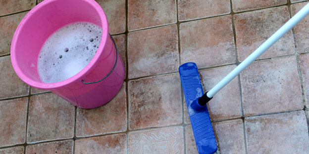 Ein pinkfarbener Eimer mit Seifenwasser und ein blauer Schrubber stehen auf einem Fließenboden.