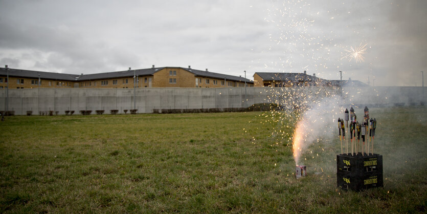 Im Hintergrund ist hinter Stacheldraht die JVA Rosdorf zu erkennen. Im Vordergrund stehen Raketen in einer Getränkekiste und es kommt Feuerwerk aus einer Feuerwerksbatterie