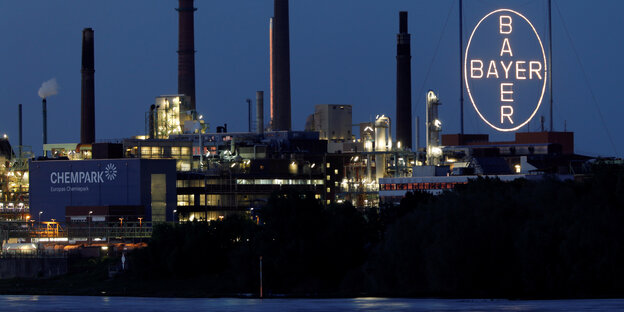Ein Chemiepark von Bayer bei Nacht. Im Hintergrund die Schlote der Fabriken, vorne rechts das Leuchtende Bayer-Emblem