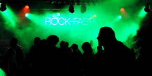 Menschen stehen auf einer Tanzfläche, die in grünes Licht und Nebel getaucht ist. Im Hintergrund leuchten die Worte "Rock-Fabrik" an der Wand.