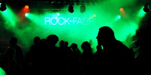 Menschen stehen auf einer Tanzfläche, die in grünes Licht und Nebel getaucht ist. Im Hintergrund leuchten die Worte "Rock-Fabrik" an der Wand.