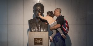 Ein kleines Mädchen auf dem Arm ihres Vaters neben ein Büste von Yitzhak Rabin