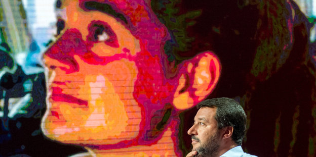 Salvini sitzt vor einem Bildschirm, auf dem ein Bild von Carola Rackete zu sehen ist
