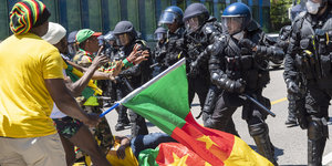 Schwarze mit der kamerunischen Fahne stehen behelmten Polizisten gegenüber. Ein Demonstrant liegt am Boden