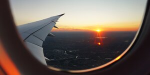 Blick vom Inneren eines Flugzeugs auf die Tragfläche. Im Hintergrund ein Sonnenuntergang