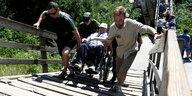 Zwei Männer helfen einem Rollstuhlfahrer dabei eine beschädigte Brücke zu überqueren