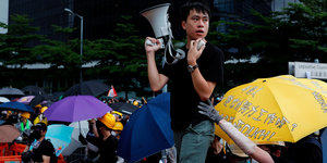 Mann mit Megafon vor aufgespannten Regenschirmen von Demonstrierenden