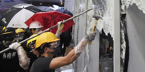 Menschen mit Regenschirmen und Bauhelmen hacken auf eine Glastür ein