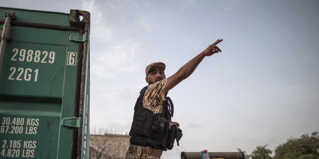 Ein Kämpfer in Camouflagehemd und schwarzer Schussweste steht neben einem dunkelgrünen Container und zeigt in die Luft.