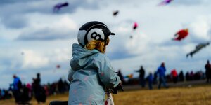 Ein Kind auf dem Tempelhofer Feld, es trägt einen Helm, hinter ihm steigen bunte Drachen