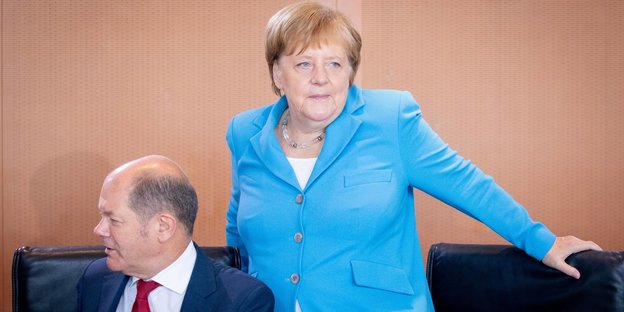 Merkel steht neben Olaf Scholz, der sitzt
