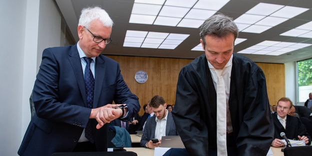 Geschäftsführer der Deutschen Umwelthilfe Jürgen Resch steht in einem Gerichtssaal neben seinem Verteidiger in Robe. Resch schaut auf seine Armbanduhr.