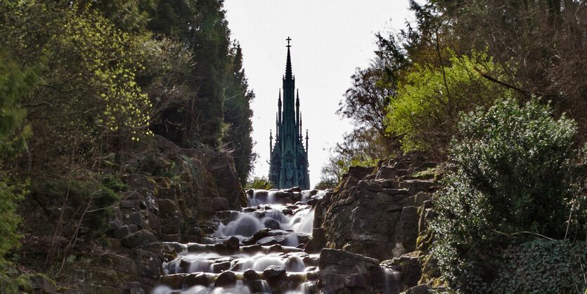 Rechts und links stehen Bäume und Büsche, dazwischen fließt ein kleiner Wasserfall. Im HIntergund die Spitze einer Skulptur.