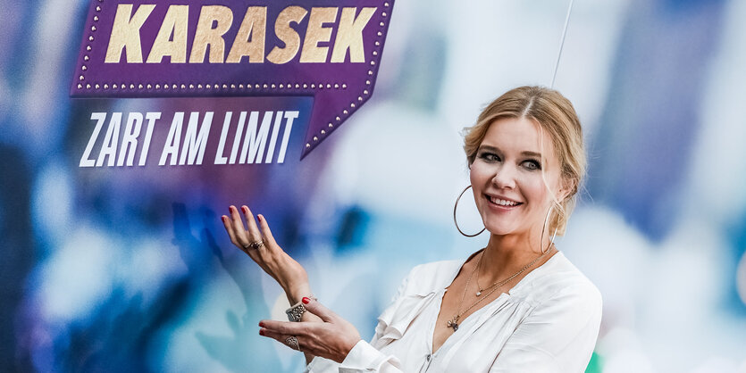 Laura Karasek steht vor dem Logo ihrer Show und macht eine präsentierende Geste