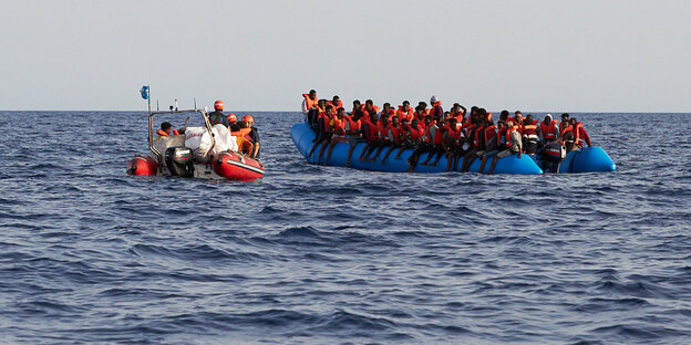 Geflüchtete auf einem völlig überfüllten Schlauchboot, daneben ein kleineres Schlauchboot mit Seenothelfern