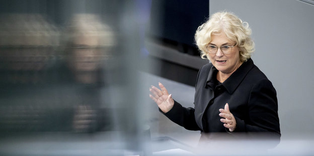 Christine Lambrecht am Rednerpult des Bundestags. Sie wird unscharf durch eine Glaswand gespiegelt