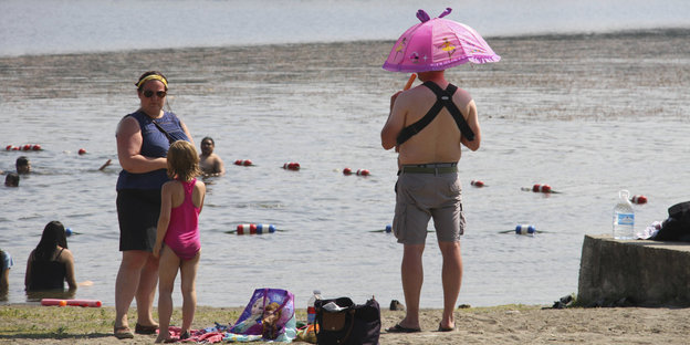 Menschen mit wenig Bekleidung am Ufer eines Gewässers. Ein Mann hält einen rosa Sonnenschirm über seinen Kopf