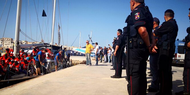 Links das Segelschiff Alex, das anlandet. Rechts am Ufer steht bereits aufgereiht die italienische Polizei