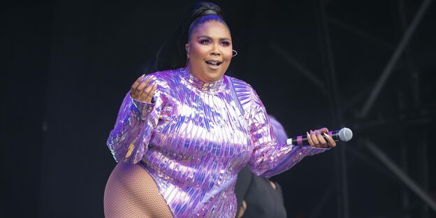 Eine Frau in lila glitzernem Bodysuit steht schnipsend auf einer Bühne Eine Frau in lila glitzernem Bodysuit steht schnipsend auf einer Bühne