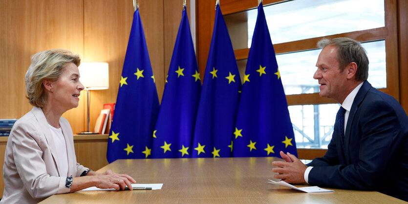 Ursula von der Leyen und Donald Tusk sitzen sich gegenüber, im Hintergrund EU-Flaggen