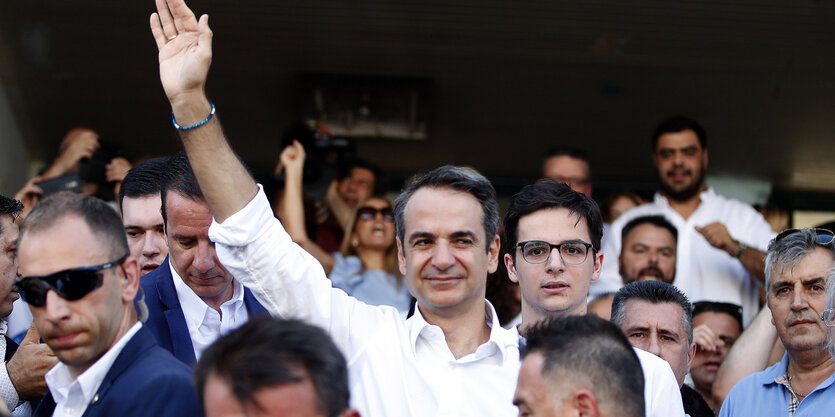 Mitsotakis steht in einer Menge und hebt schmunelnd den Arm