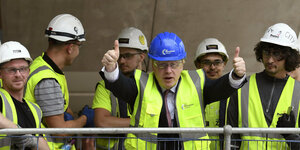 Boris Johnson steht mit Bauarbeiter-Helm und -Weste umringt von Bauarbeitern
