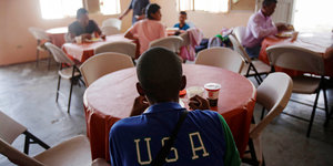 Ein Mann sitzt an einem Tisch in einer Unterkunft für Geflüchtete in Mexiko. Er tägt ein T-Shirt mit der Aufschtift USA