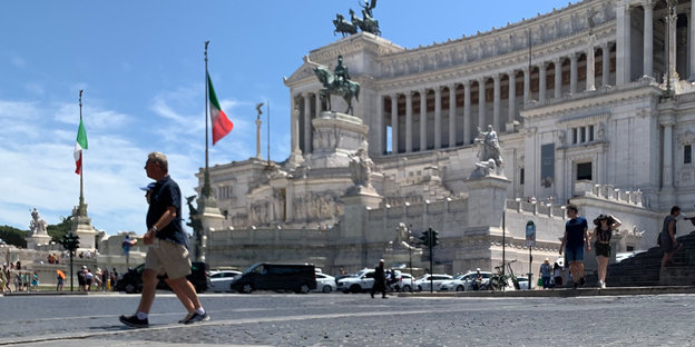 Passanten gehen vor dem Denkmal für König Vittorio Emanuele II. in Rom über Kopfsteinpflaster.