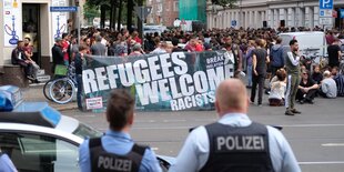 Teilnehmer einer Demonstration versammeln sich auf einer Straße. Sie haben ein Transparent auf dem refugees Welcome steht. Die Polizei beobachtet sie.