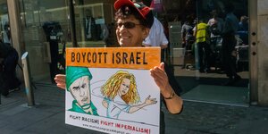 Eine Frau mit Cappy und Sonnenbrille steht auf einem Brügerstreig vor einer Ladenzeile und hält ein Schild in beiden Händen. Daraif steht "Boycott Israel".