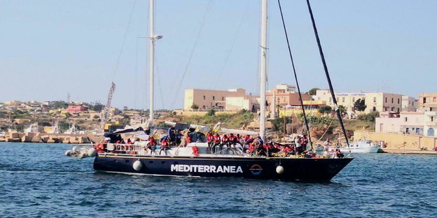 ein mit Flüchtlingen gefülltes Segelschiff vor einem Hafen