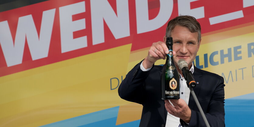 ein Mann steht vor einem Mikrofon und hält eine Flasche Sekt in den Händen